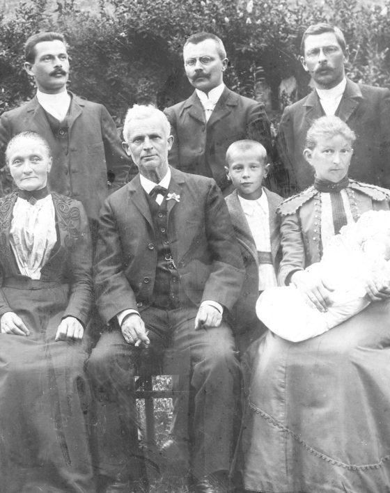 Altes Schwarz-Weiß-Foto mit Männern in dunklen Anzügen, Frauen in langen Kleidern und Kindern.
