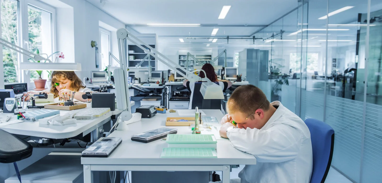 Drei Uhrmacher bei der Arbeit an Uhrmachertischen.