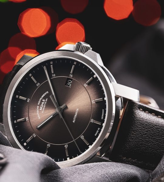 Armbanduhr 29er Casual mit samtbraunem Zifferblatt am braunen Lederband liegt seitlich auf braun-grauem Hemd. Dahinter kleine orange-rote Lichtpunkte.