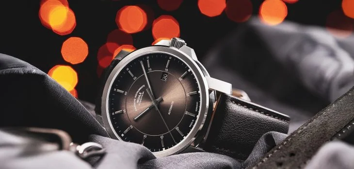 Armbanduhr 29er Casual mit samtbraunem Zifferblatt am braunen Lederband liegt seitlich auf braun-grauem Hemd. Dahinter kleine orange-rote Lichtpunkte.