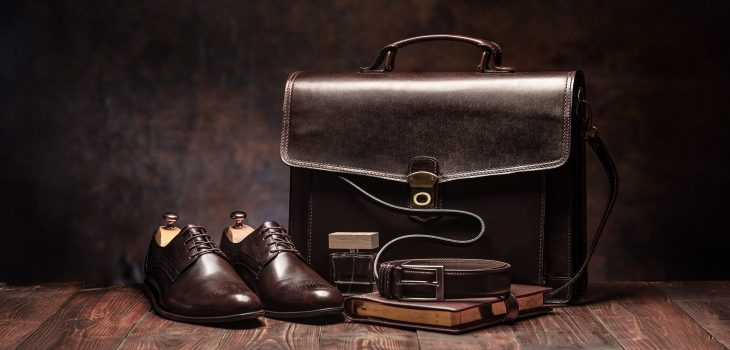 Braune Leder-Aktentasche, braune Lederschuhe, ein Notizbuch und ein brauner Gürtel auf einem Holzboden mit braunem Hintergrund.