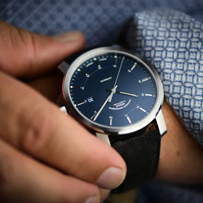Wristwatch (29er Casual) with blue dial, silver indices and case on a dark leather strap on a wrist with a blue patterned shirt sleeve.