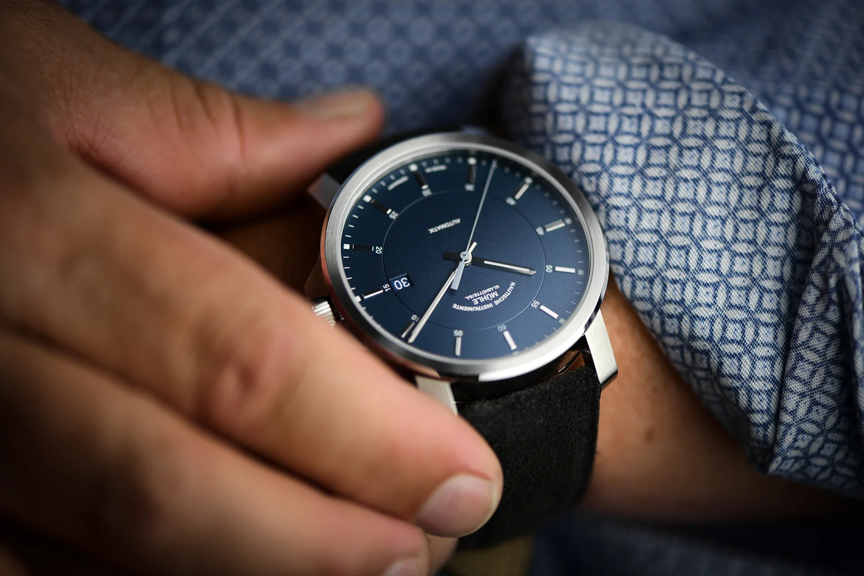 Armbanduhr (29er Casual) mit blauem Zifferblatt, silbernen Indizes und Gehäuse am dunklen Lederband an einem Handgelenk mit blau-gemustertem Hemdsärmel.