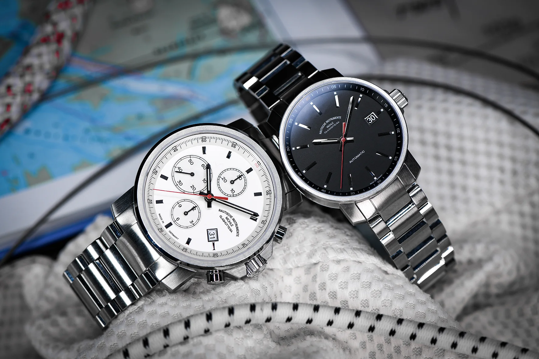 Zwei Armbanduhren (links: 29er Chronograph mit weißem Zifferblatt, recht: 29er mit schwarzem Zifferblatt) mit Metallbändern liegen auf einer weißen Jacke und einer Landkarte.