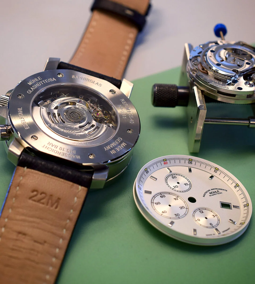 Eine Armbanduhr, ein Zifferblatt und ein Uhrwerk liegen auf einer grünen Tischunterlage.