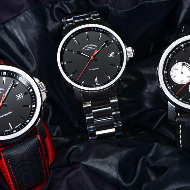 Drei Armbanduhren mit schwarzen Zifferblättern (links am rot-schwarzen Leder-Kautschukband, Mitte am Metallband, rechts am schwarzen Lederband) stehen auf einer schwarzen Lederjacke.