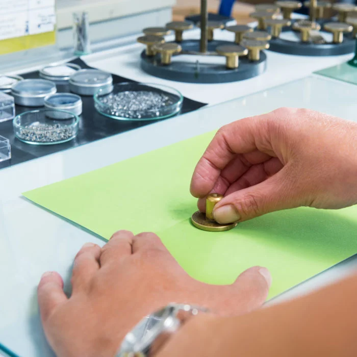 Hände einer Uhrmacherin halten ein goldenes Metallstück auf ein grünes Stück Schleifpapier. Im Hintergrund Teile und Werkzeuge.