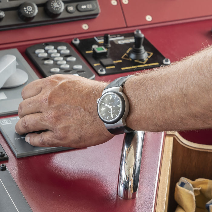 Eine schwarze Armbanduhr (S.A.R. Rescue-Timer) an einem Handgelenk vor den Navigationsgeräten der Kommandobrücke.