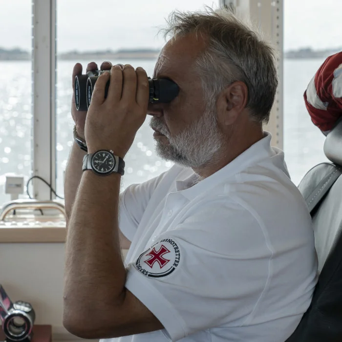 Seenotretter blickt durch ein Fernglas, an seinem Arm ein S.A.R. Rescue-Timer.