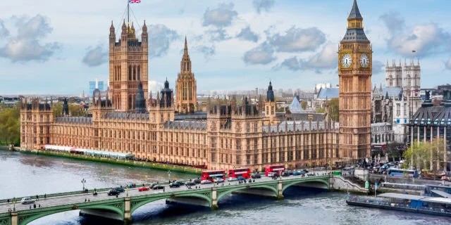 Westminster Palace mit Big Ben, im Vordergrund ein Fluss und eine Brücke. Im Hintergrund ist ein blauer Himmel mit Wolken zu sehen.