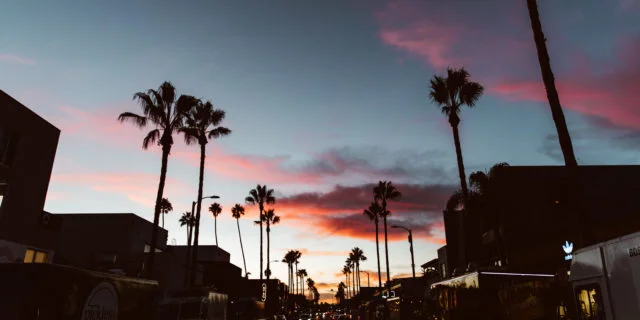 Straße in Los Angeles, die mit Palmen gesäumt ist. Im Hintergrund ein rosa-blauer Himmel mit Wolken