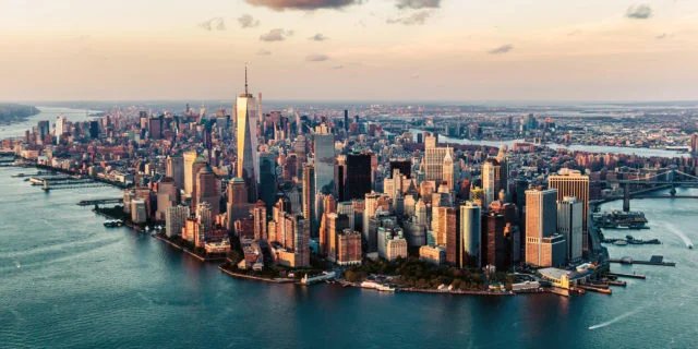Lufansicht von der Skyline von New York. Im Vordergrund fließt ein Fluss, der Himmel im Hintergrund ist leicht bewölkt.