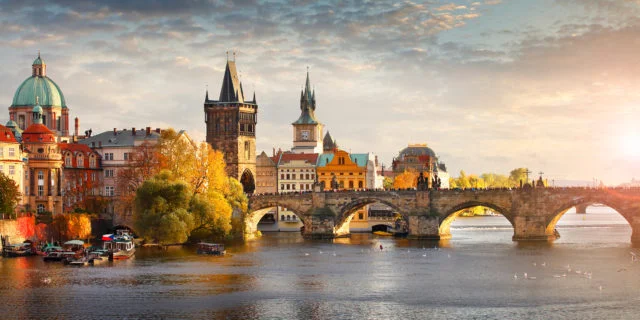 Blick auf die Karlsbrücke in Prag. Im Vordergrund fließt die Moldau. Im Hintergrund sind Gebäude und ein wolkiger Himmel zu sehen.