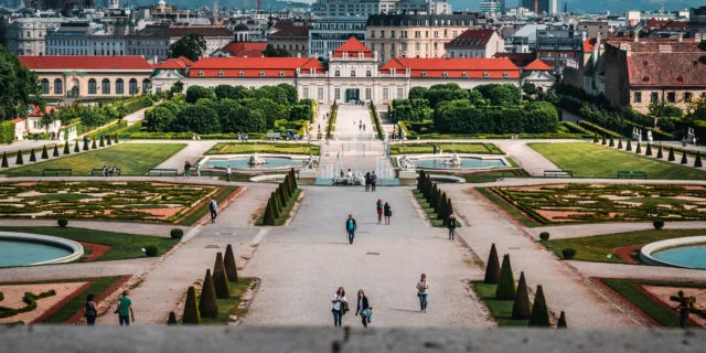 Im Vordergrund ein symmetrisch angelegter Park. Im Hintergrund ein langgezogenes Gebäude, dahinter weitere Häuser.