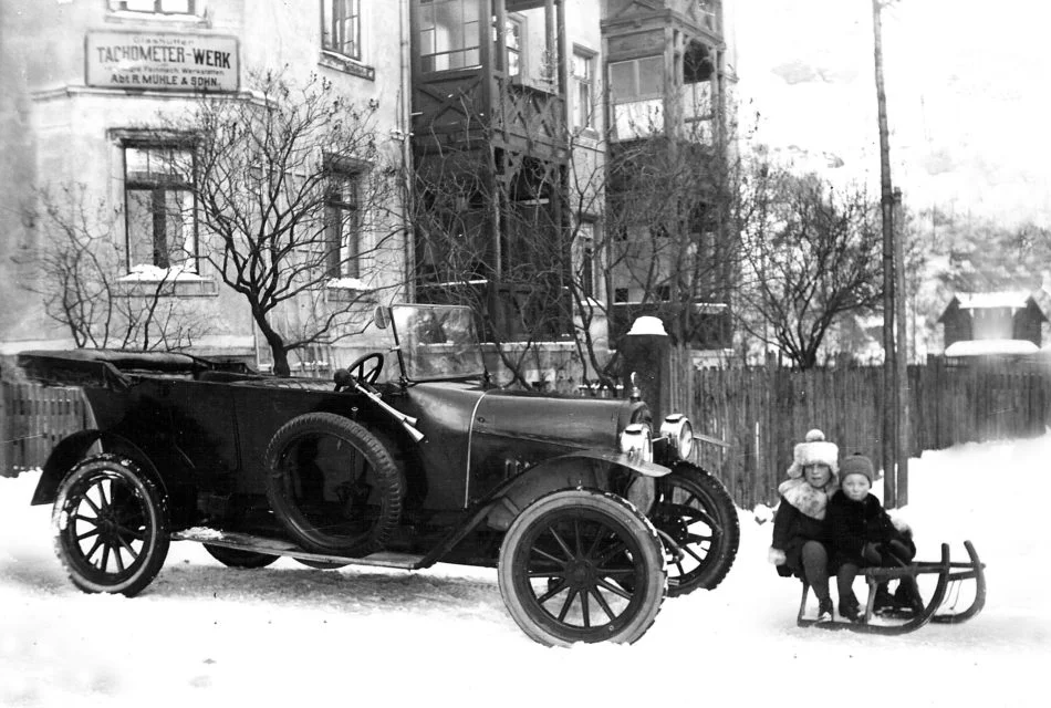 Altes Schwarz-Weiß-Foto mit einem Oldtimer und zwei Kindern auf einem Schlitten vor dem Stammsitz von Mühle-Glashütte.