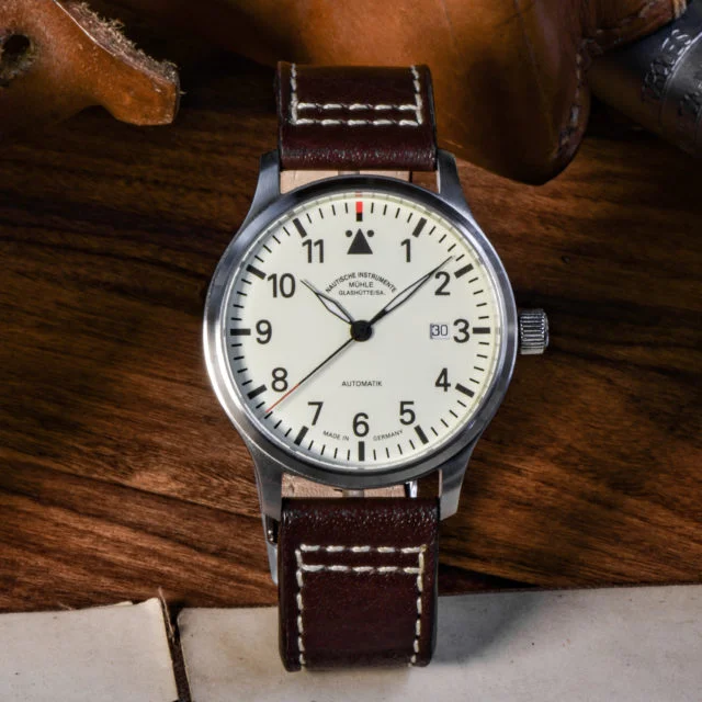 Terrasport II: Armbanduhr mit cremefarbenem Zifferblatt, schwarzen Indizes und silbernem Gehäuse am braunen Lederband steht auf einem Holztisch mit Papieren und Lederstücken.