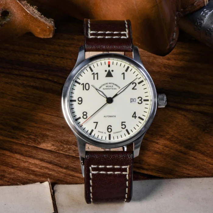 Terrasport II: Armbanduhr mit cremefarbenem Zifferblatt, schwarzen Indizes und silbernem Gehäuse am braunen Lederband steht auf einem Holztisch mit Papieren und Lederstücken.