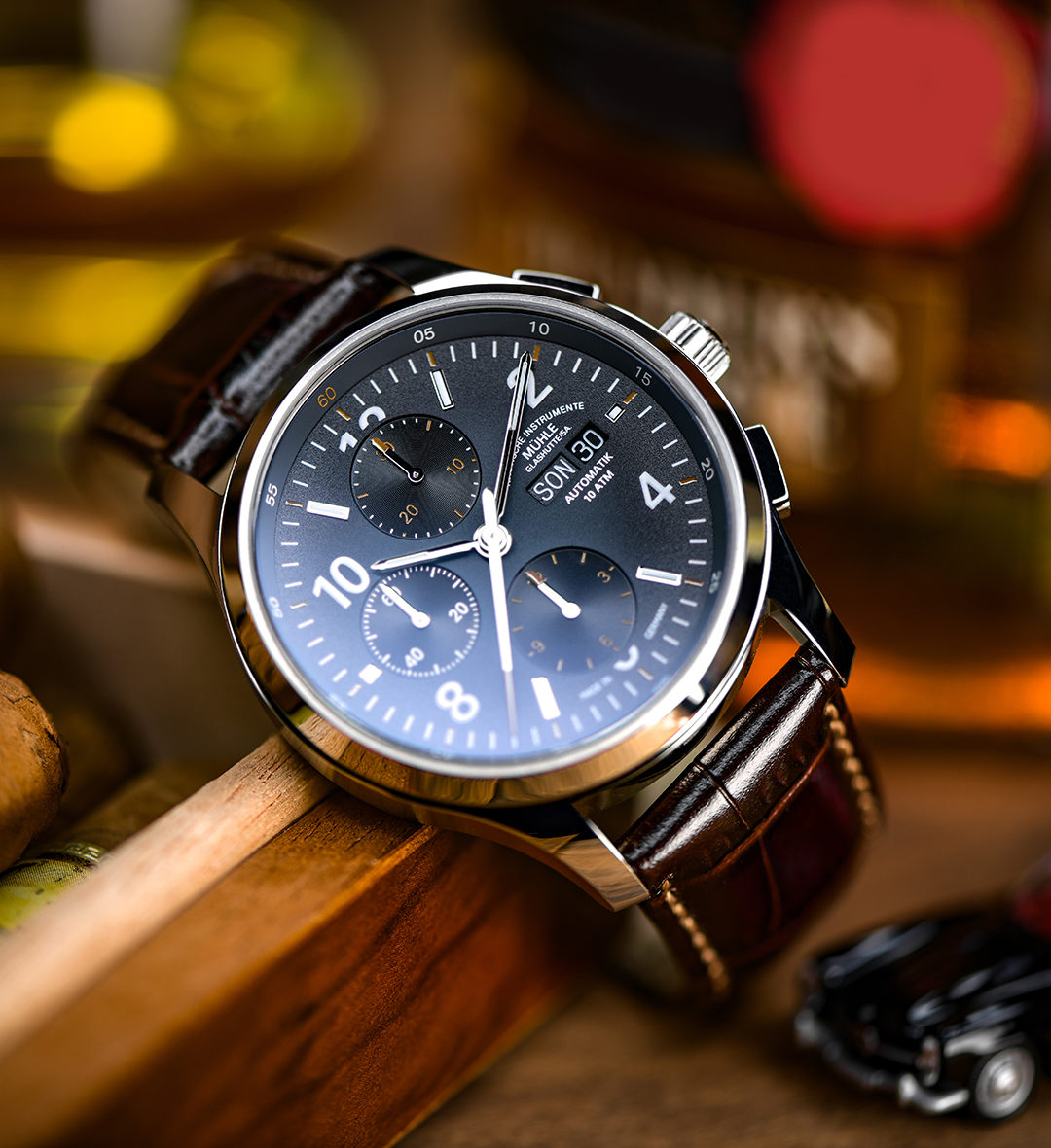 A Lunova Chronograph wristwatch with a brown leather strap stands slightly askew on a wooden table, with a black toy car in front of it.
