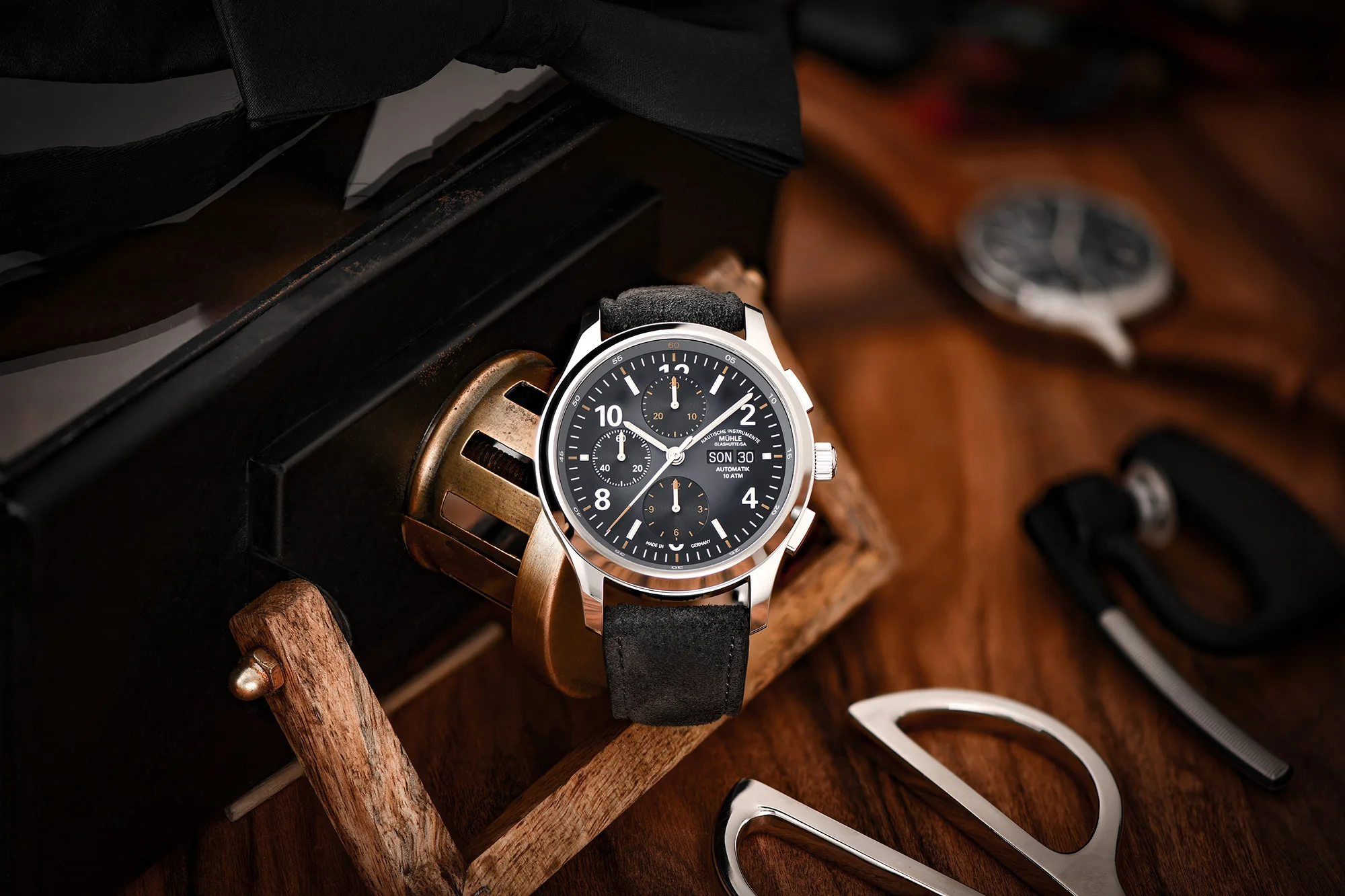 Armbanduhr Lunova Chronograph am grauen Veloursband steht auf einem Holztisch, daneben liegt eine schwarze Kiste, eine Schere und eine weitere Uhr.