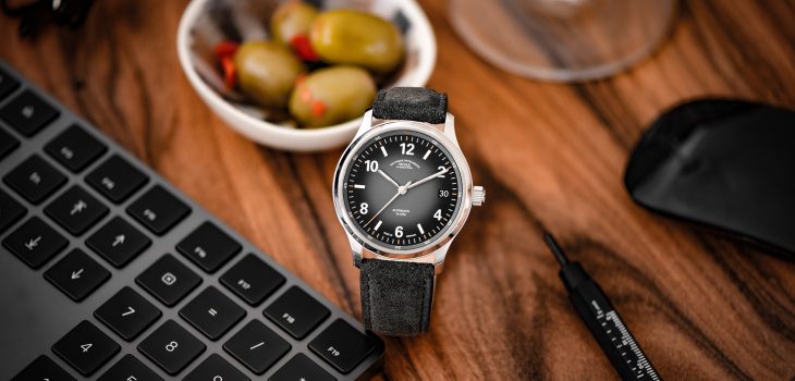 Armbanduhr Lunova Datum am grauen Veloursband steht auf einem Holztisch neben einer Tastatur und einem Schälchen mit Oliven.