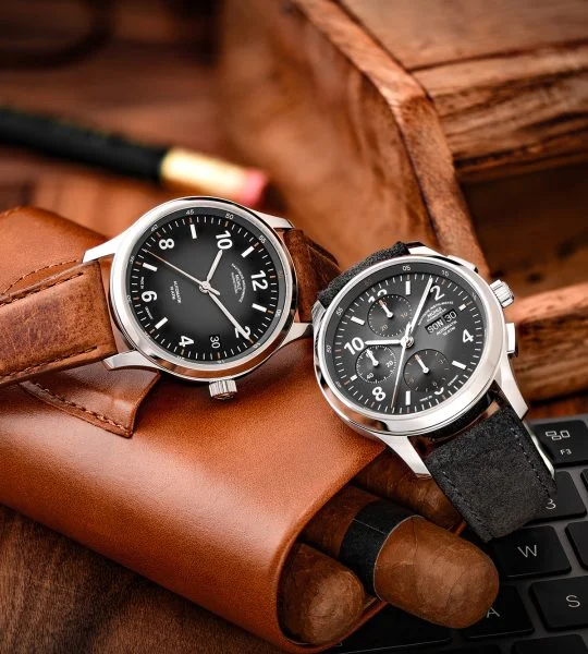 Zwei Armbanduhren (Lunova Datum am braunen Lederband, Lunova Chronograph am grauen Veloursband) liegen auf einem braunen Lederetui mit Zigarren.