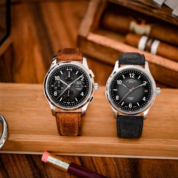 Zwei Armbanduhren (Lunova Chronograph am braunen Lederband, Lunova Datum am grauen Veloursband) stehen auf einem Holzbrett. Im Hintergrund zwei Zigarren.