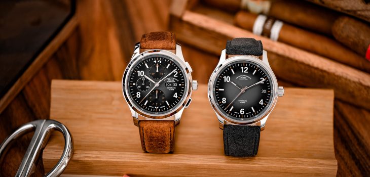 Zwei Armbanduhren (Lunova Chronograph am braunen Lederband, Lunova Datum am grauen Veloursband) stehen auf einem Holzbrett. Im Hintergrund zwei Zigarren.