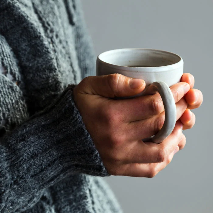 Hände einer Person mit grauem Pullover, die eine weiße Kaffeetasse halten.