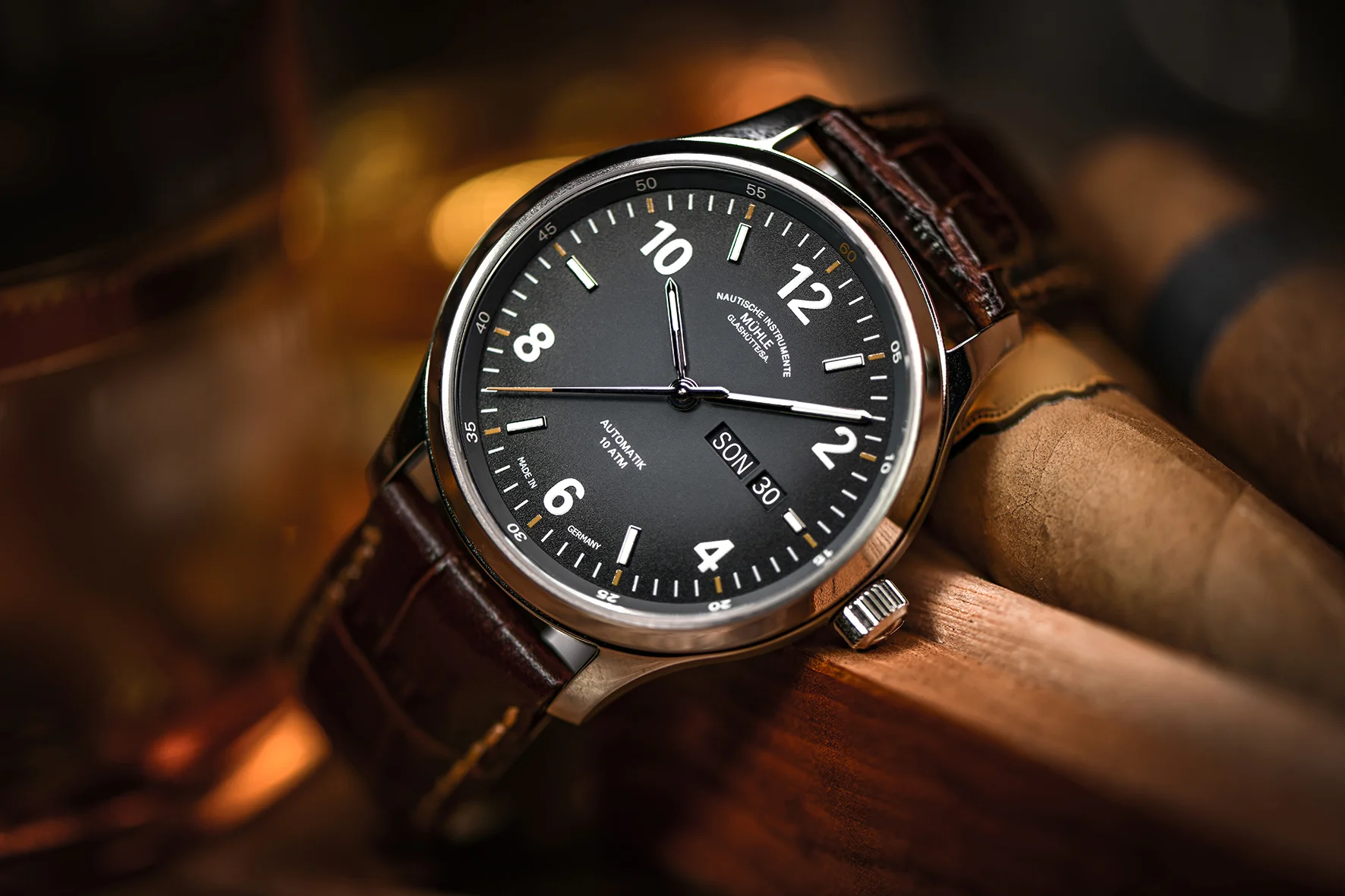 Armbanduhr mit dunkelgrauem Zifferblatt, weißen Indizes, silbernem Gehäuse und braunem Lederband vor gerollten Zigarren.