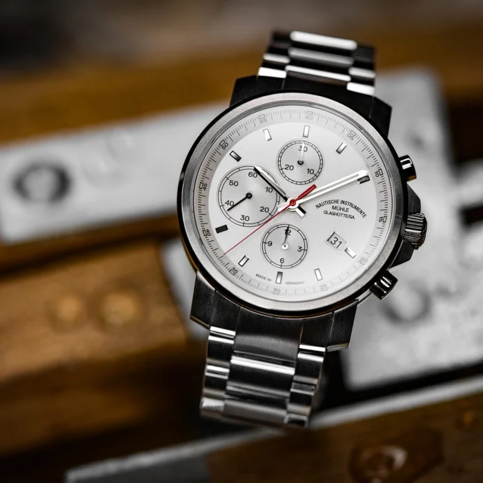 Armbanduhr "29er Chronograph" mit weißem Zifferblatt und Metallband steht auf einem Holzuntergrund mit Metallstücken.