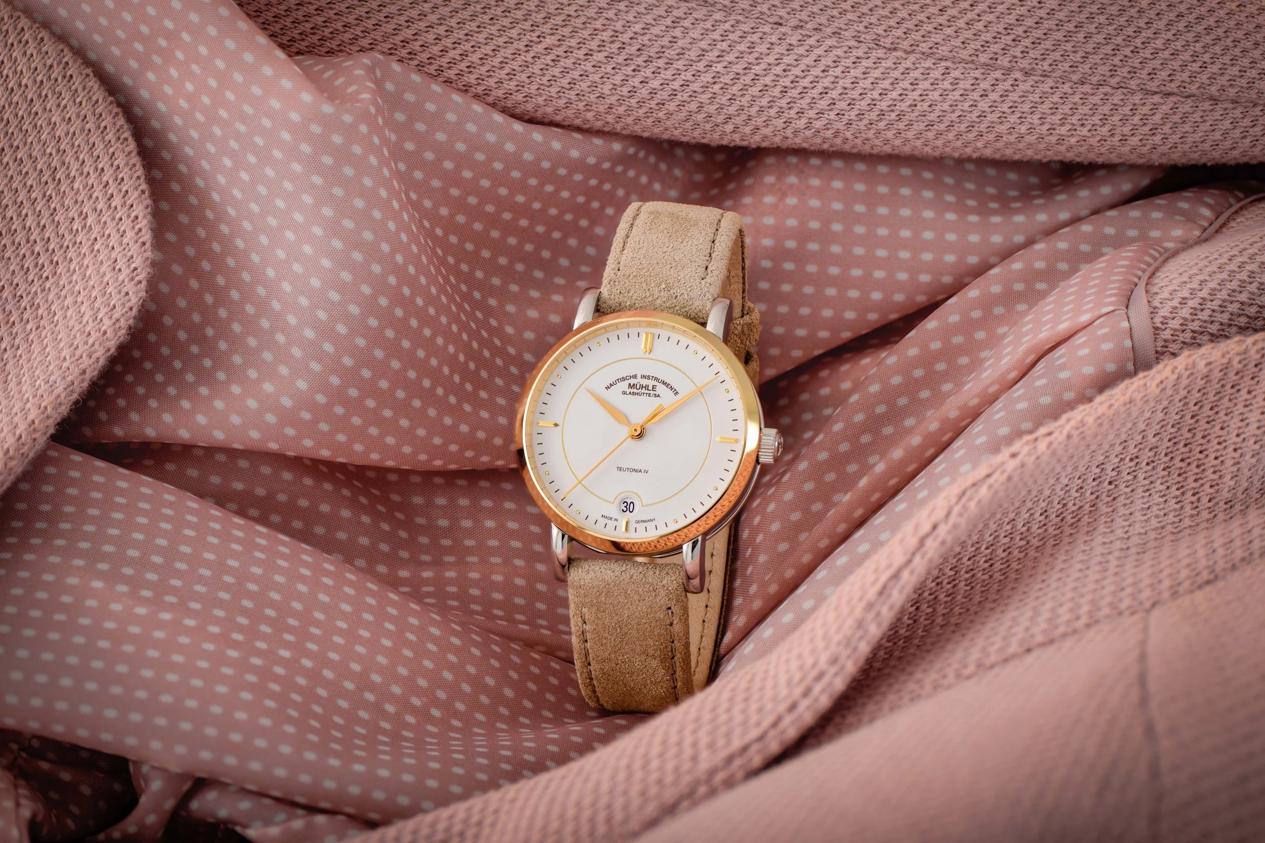 Armbanduhr Teutonia IV Lady mit weißem Zifferblatt am beigen Veloursband steht auf rosa-gemustertem Stoff.