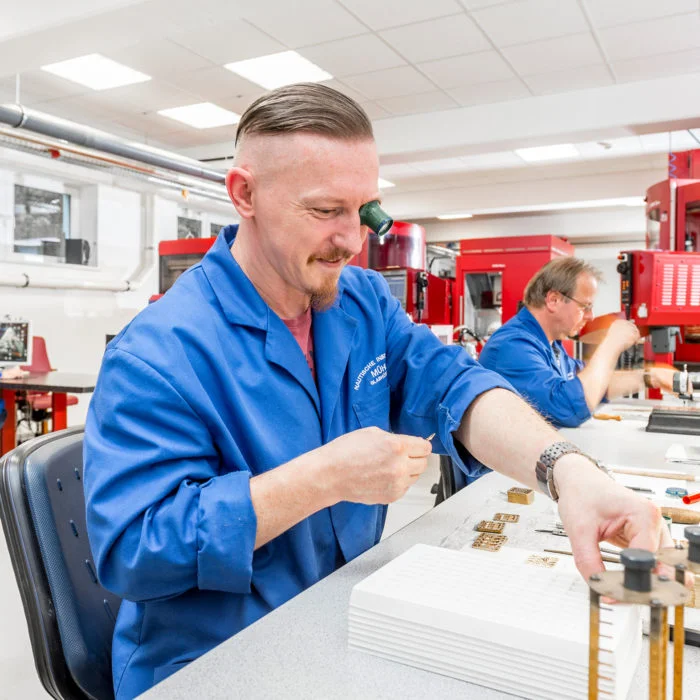 2 Mitarbeiter der CNC-Fertigung mit blauen Kitteln arbeiten an ihrem Werktisch.