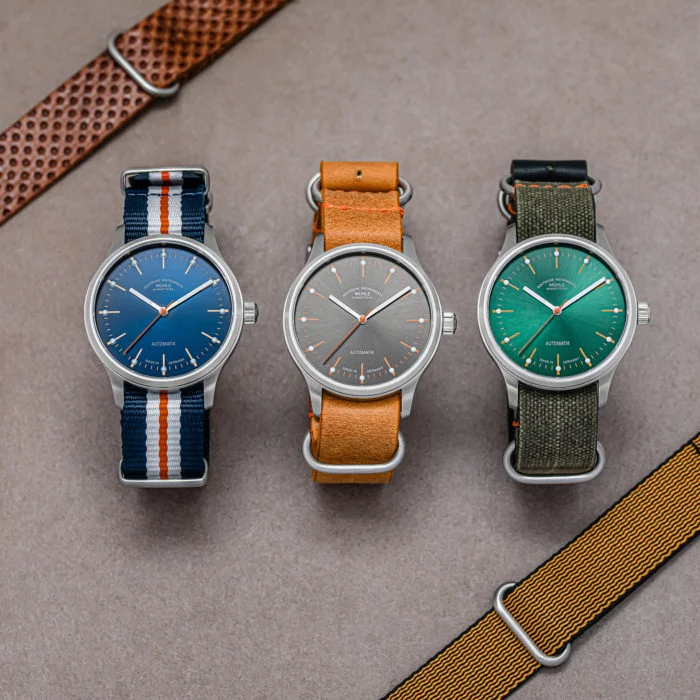 Drei Armbanduhren (Panova blau, grau und grün) liegen mit zwei zusätzlichen Bändern auf einem grauen Tisch.