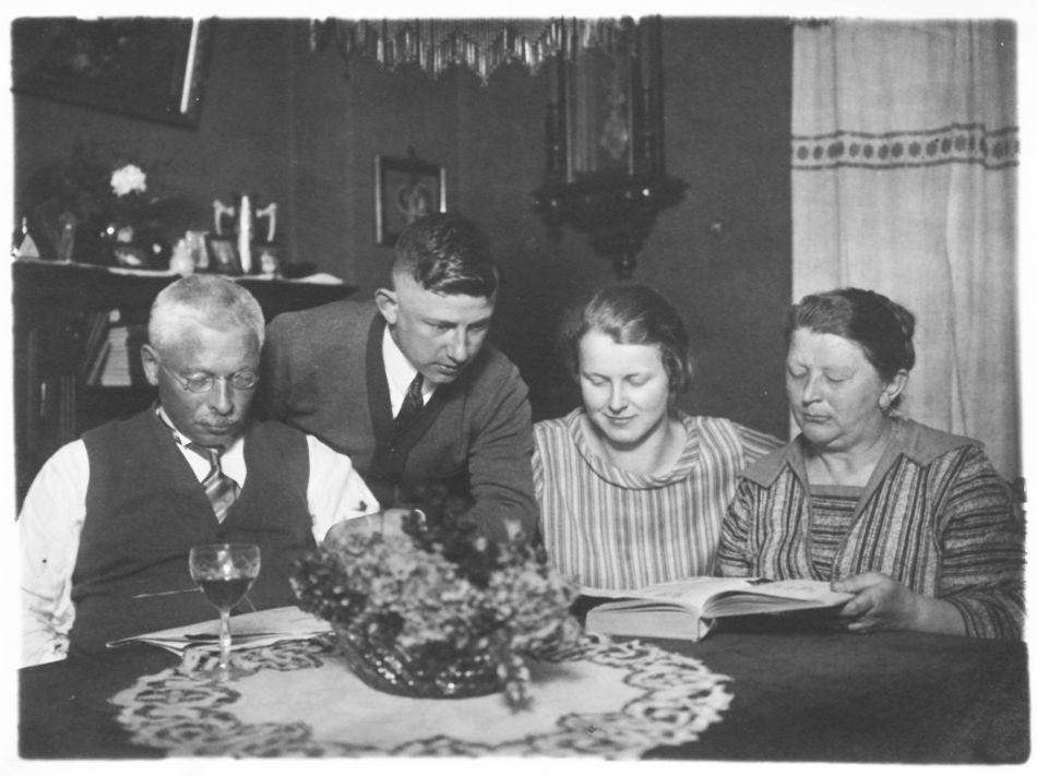 Altes Schwarz-Weiß-Foto mit zwei Männern und zwei Frauen an einem Tisch mit Blumen und Tischdecke.