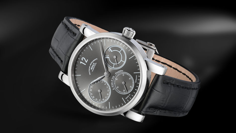 Liegende Armbanduhr mit dunkelgrauem Zifferblatt, drei Hilfszifferblättern, silbernem Gehäuse und schwarzem Lederband vor schwarzem Hintergrund.