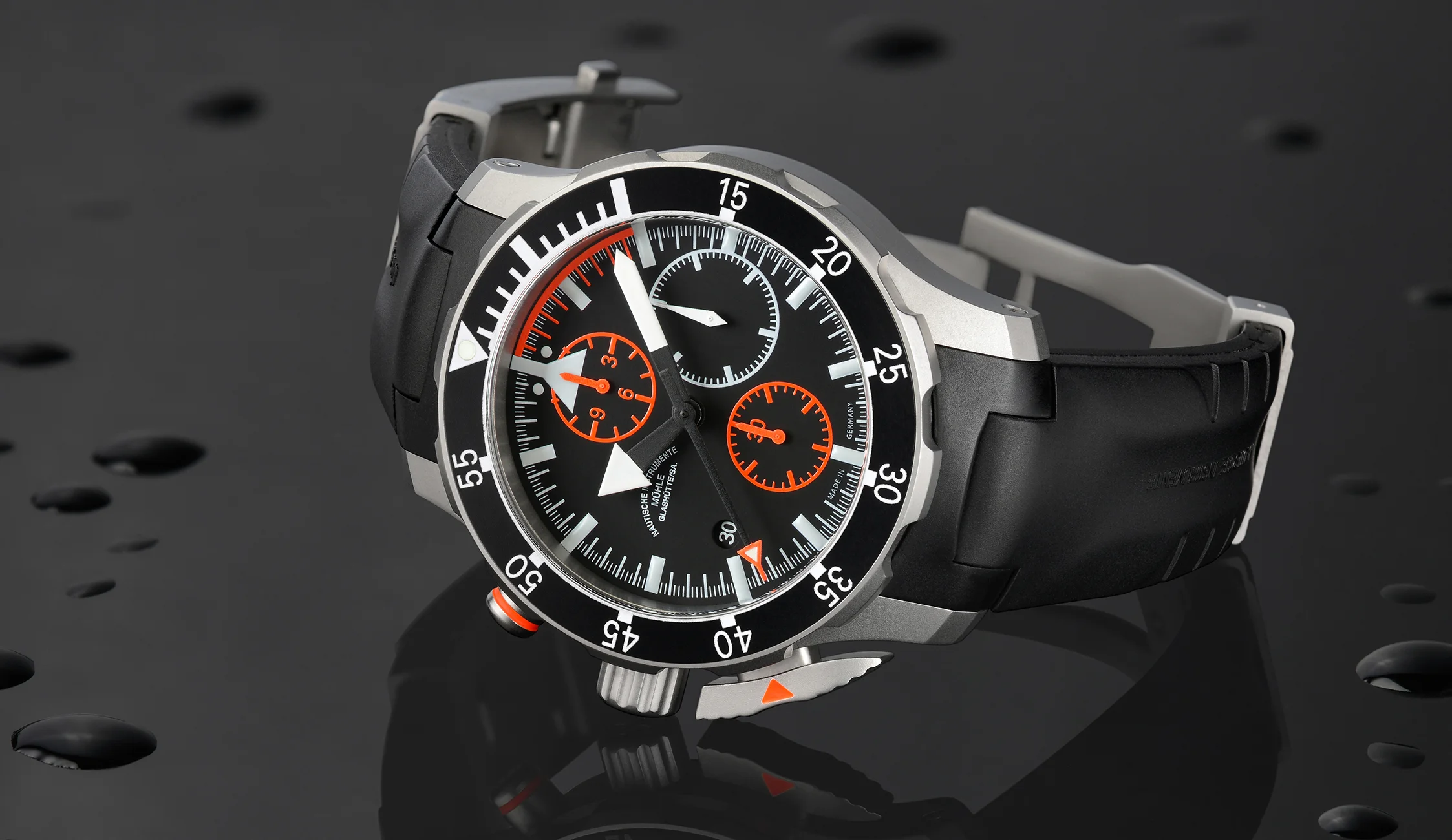 S.A.R. Fliegerchronograph – eine Armbanduhr mit schwarzem Zifferblatt und roten Details, schwarzer Lünette, mattsilbernem Gehäuse und schwarzem Kautschukband – liegt auf einer spiegelnden Fläche mit Wassertropfen.