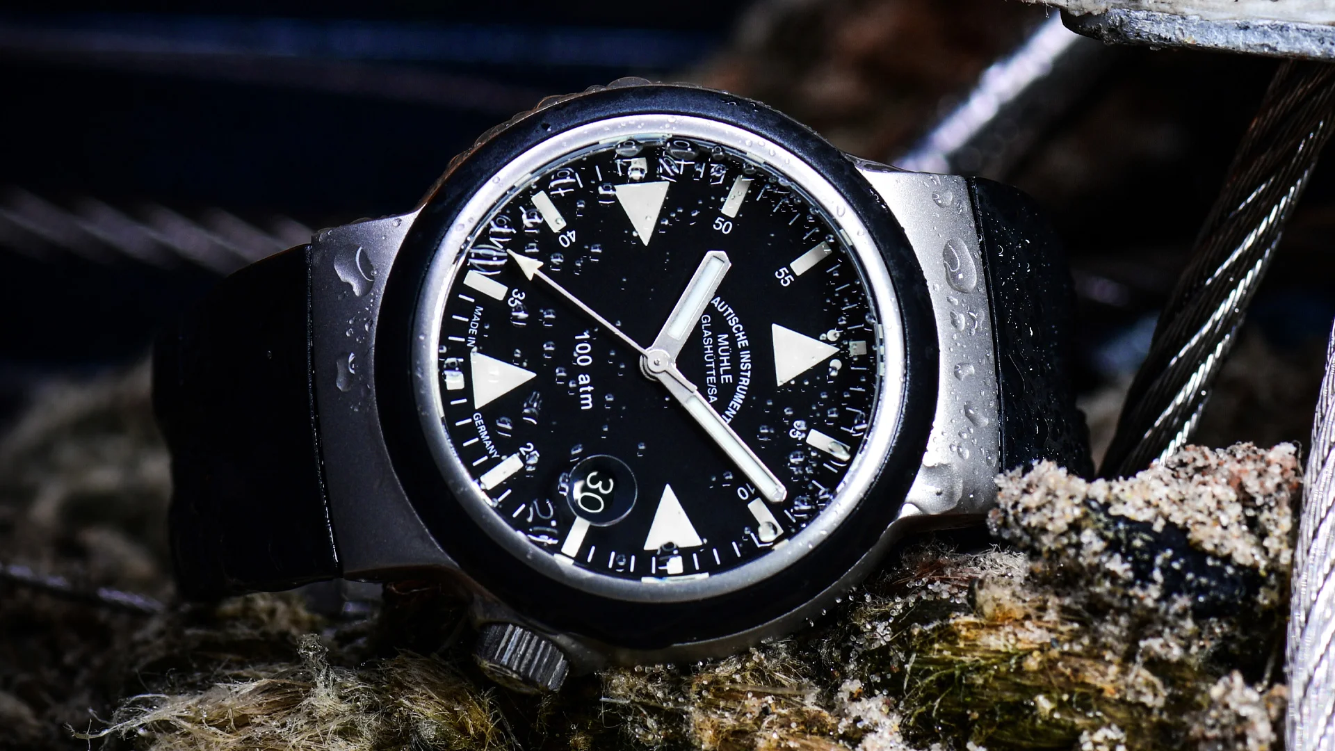 Eine Armbanduhr mit schwarzem Zifferblatt, weißen Zeigern und mattsilbernem Gehäuse liegt auf einem Stein vor Tauen.