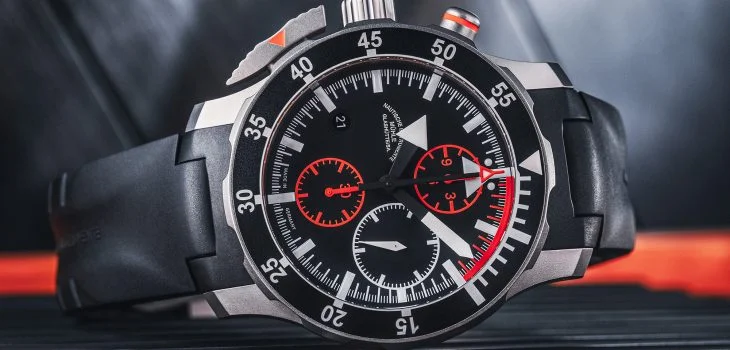 S.A.R. Fliegerchronograph am schwarzen Kautschukband liegt seitlich auf einer orange-grauen Oberfläche.