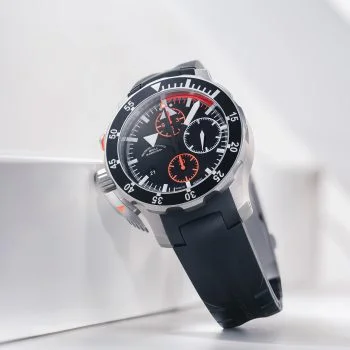 S.A.R. Fliegerchronograph am schwarzen Kautschukband steht auf einem weißen Tisch mit einem weißen Kasten dahinter.