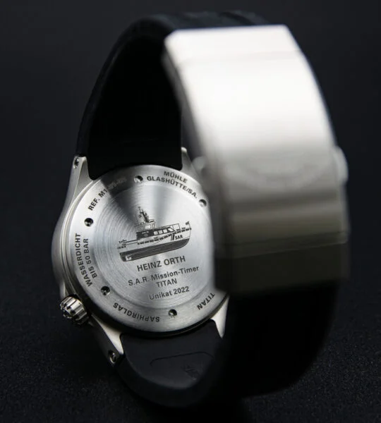 Eine Armbanduhr liegt mit dem Zifferblatt nach unten auf einer schwarzen Fläche. Der Gehäuseboden zeigt einen S.A.R. Rettungskreuzer. Im Vordergrund eine silberfarbene Schließe.