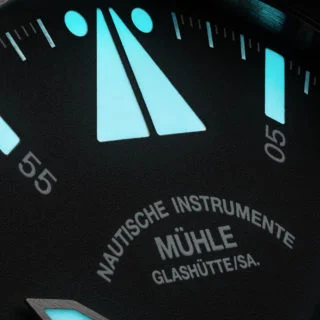 Nahaufnahme des blau-grün nachleuchtenden Fliegerdreiecks mit zwei Punkten und nachleuchtenden Strichindizes des S.A.R. Mission-Timers. Unten das Mühle-Logo des Zifferblatts.