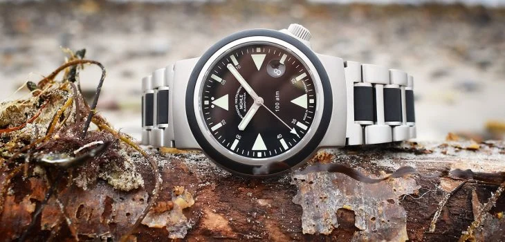 S.A.R. Rescue-Timer mit schwarzem Zifferblatt am Metall-/Kautschukband liegt seitlich auf einem Stück Holz, im Hintergrund das Meer.