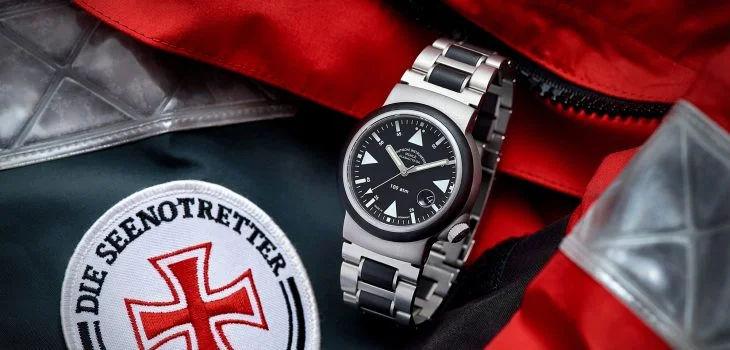 S.A.R. Rescue-Timer mit schwarzem Zifferblatt am Metall-/Kautschukband steht auf einer roten Jacke der Seenotretter mit Aufnäher der DGzRS