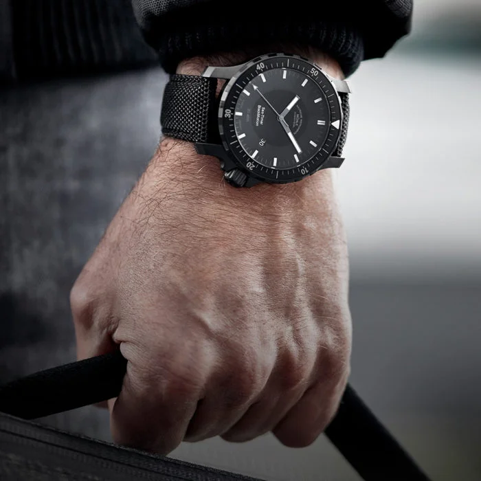 Armbanduhr "Sea-Timer BlackMotion" wird am Handgelenk getragen. Die Hand hält eine dunkle Tasche fest.