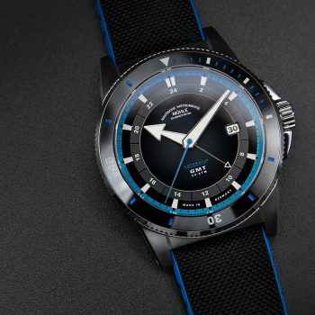 Eine Armbanduhr (Sportivo Travel GMT) mit schwarzem Zifferblatt mit blauen Details, schwarzer Lünette, silbernem Gehäuse und schwarz-blauem Band liegt auf einer schwarzen Platte.