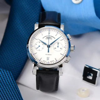 Armbanduhr Teutonia IV Chronograph am schwarzen Lederband steht auf einem weißen Tisch, auf dem eine blaue Krawatte, ein blauer Hemdsärmel und ein Manschettenknopf liegen.