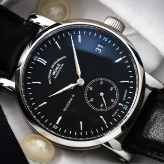 Armbanduhr "Teutonia IV Kleine Sekunde" mit schwarzem Zifferblatt und schwarzem Lederband liegt auf einem weißen Hemdsärmel.