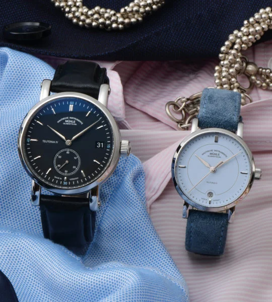 Zwei Armbanduhren (Teutonia IV Kleine Sekunde und Teutonia IV Lady mit hellblauem Zifferblatt) stehen auf blauem und rose Stoff mit Schmuck im Hintergrund.