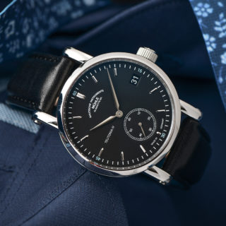 Armbanduhr "Teutonia IV Kleine Sekunde" mit schwarzem Zifferblatt und Lederband steht auf blauem Stoff.