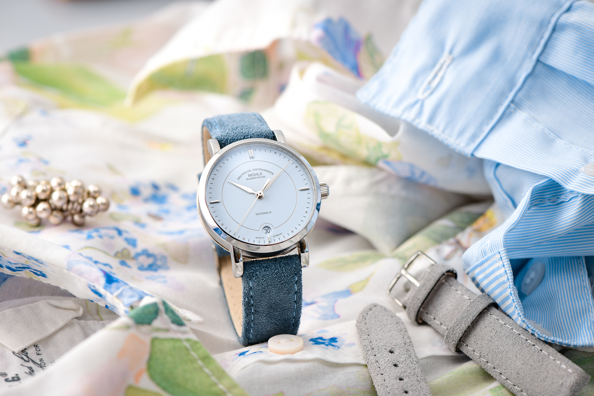 Armbanduhr Teutonia IV Lady mit pastellblauem Zifferblatt am blauen Veloursband steht einer weißen Bluse mit blauen und grünen Blümchen. Daneben ein weiteres Band und eine blaue Bluse.